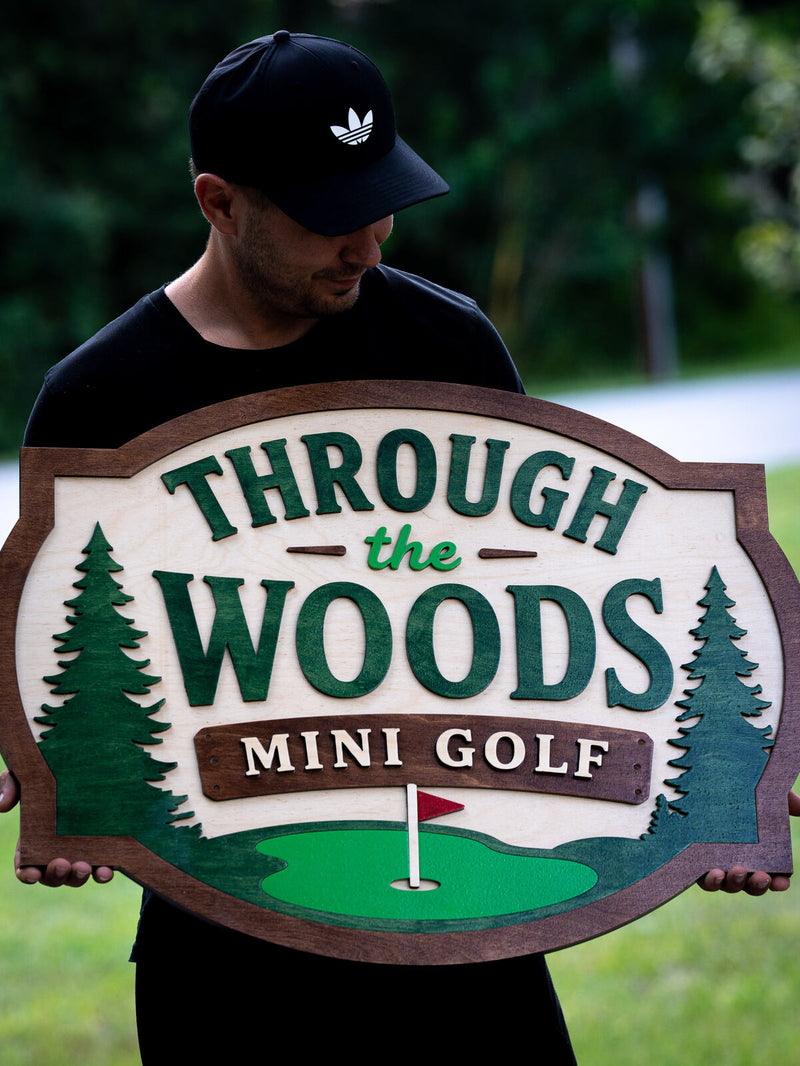 Cream shaped wood sign with a dark walnut border, layered dark green pines either side of green lettering reading Through the Woods Mini Golf
