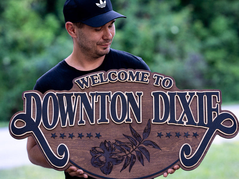 Wide scrolled warm walnut sign with navy layered lettering and cream outlines reading Welcome to Downton Dixie, with navy stars and an engraved pecan branch