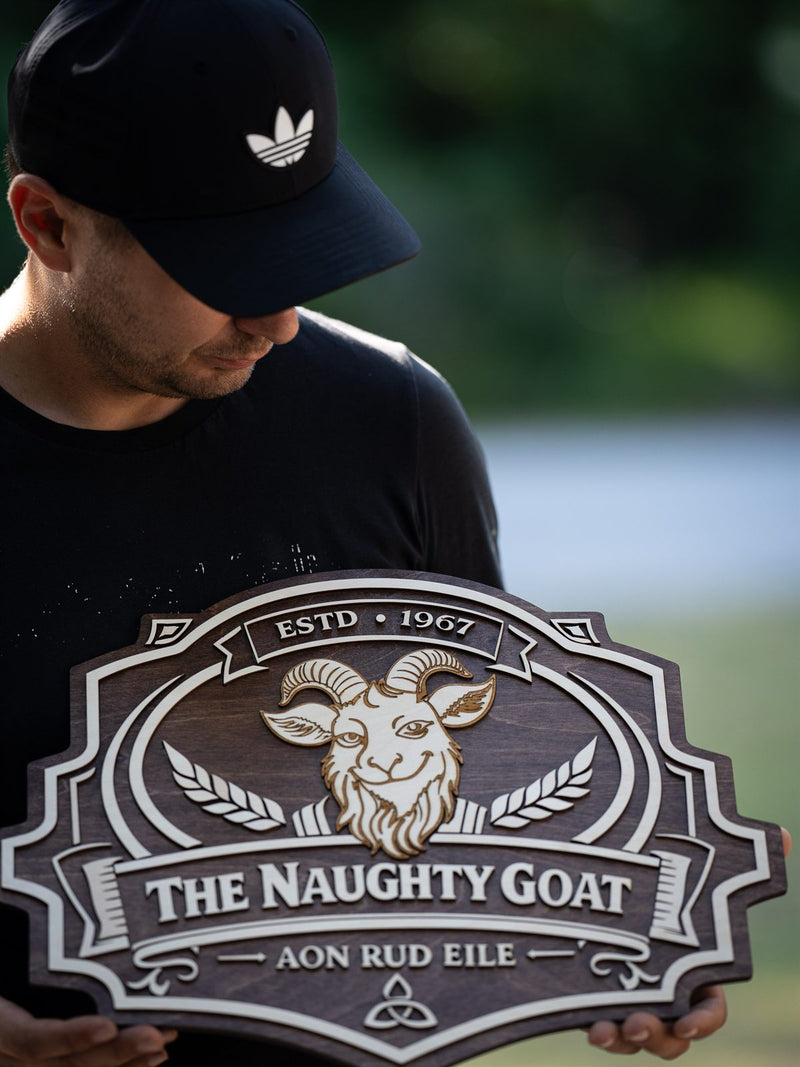 Scalloped dark walnut shield pub sign for The Naughty Goat with an engraved horned goat head, crossed barley and an Irish motto