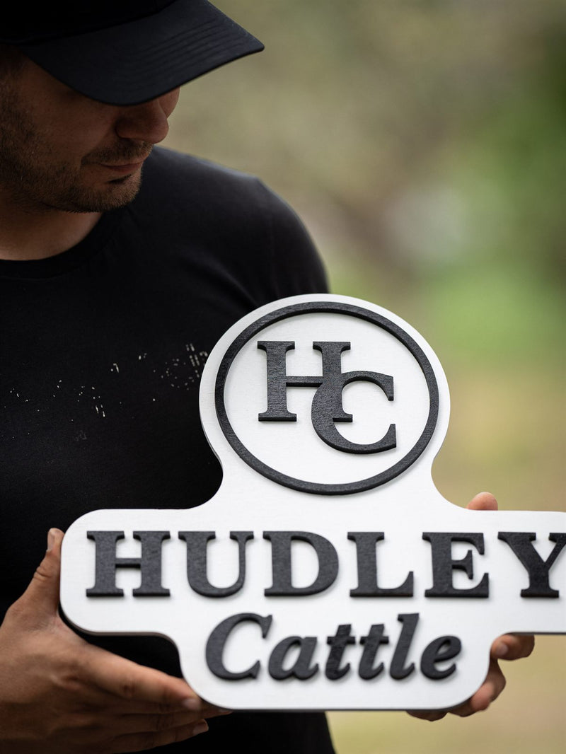 Shaped white wood ranch sign with a charcoal HC brand monogram in a circle above serif and script lettering reading Hudley Cattle
