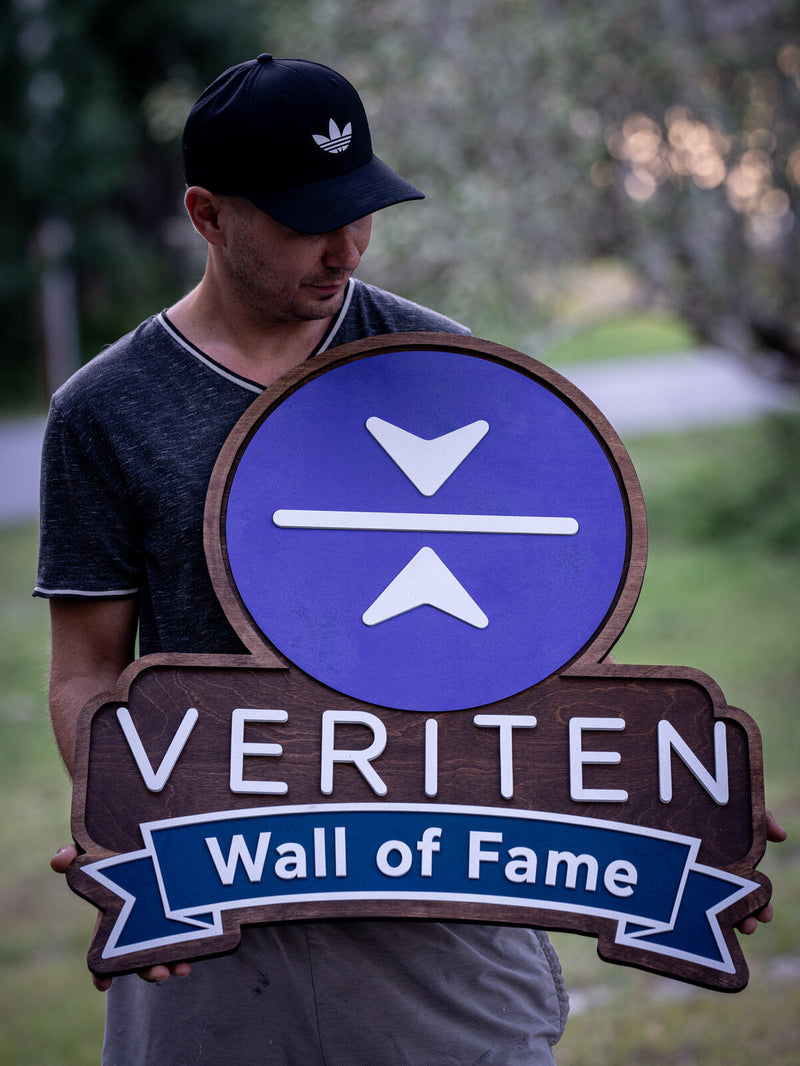 Walnut-stained shaped wood plaque with a violet disc carrying a white chevron mark, reading Veriten, Wall of Fame
