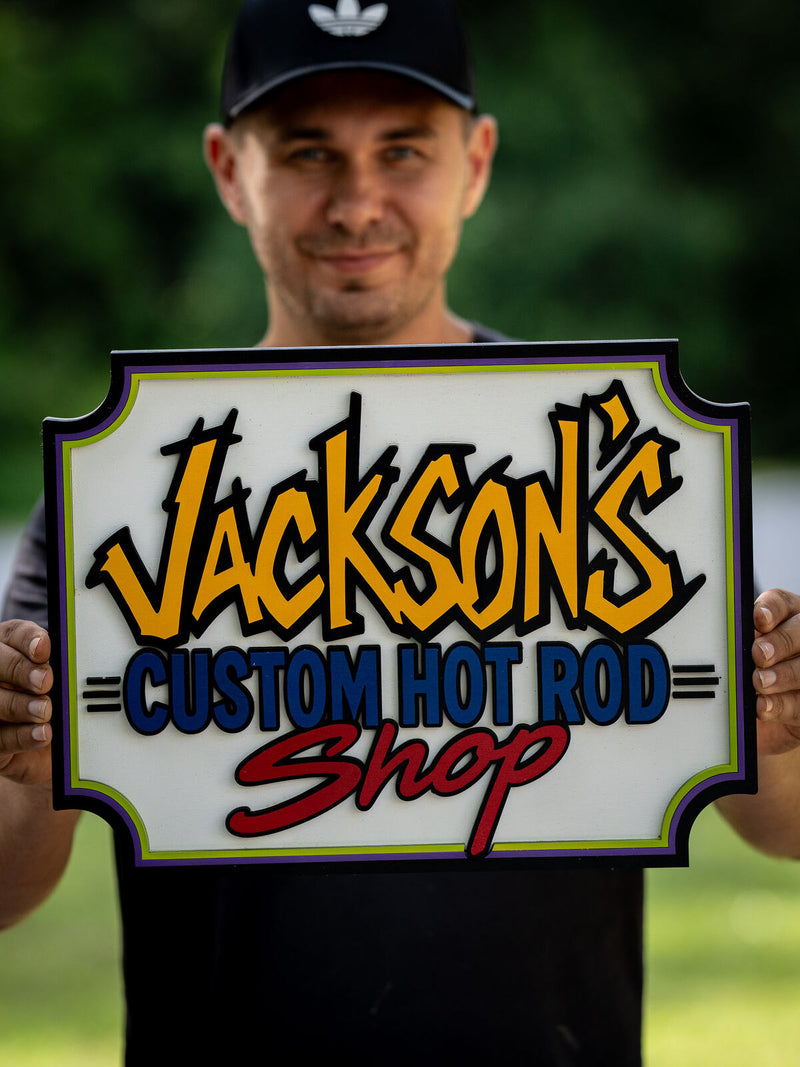 Rectangular cream wood shop sign with lime green, purple and black stacked borders and yellow-orange graffiti style lettering reading Jackson's Custom Hot Rod Shop