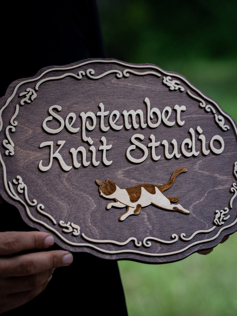 Oval mauve-brown wood studio sign with a scalloped scrolled cream border, cream script lettering and a small calico cat