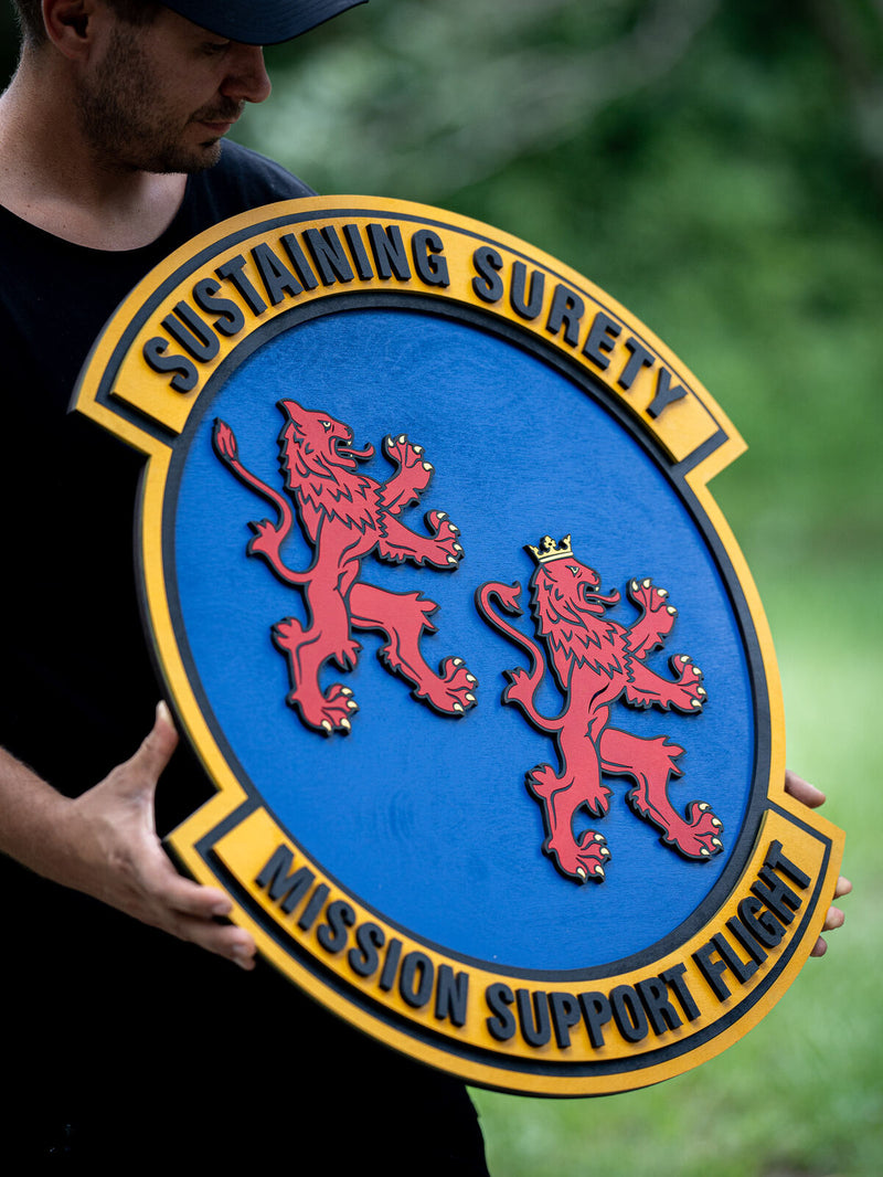 Large blue oval wood unit crest with two red rampant lions, one crowned in gold, inside a golden yellow scroll border with black motto lettering