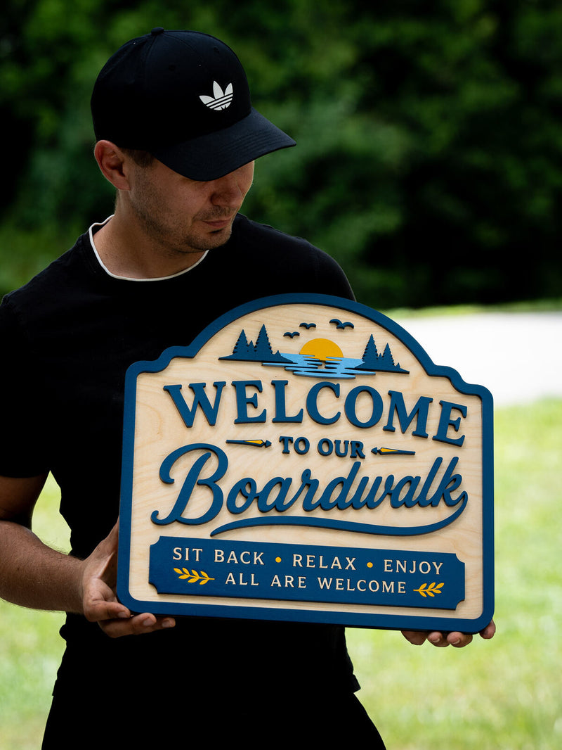 Arched cottage-shaped pale birch welcome sign with a teal border, a blue lake scene with pines, gulls and a setting sun